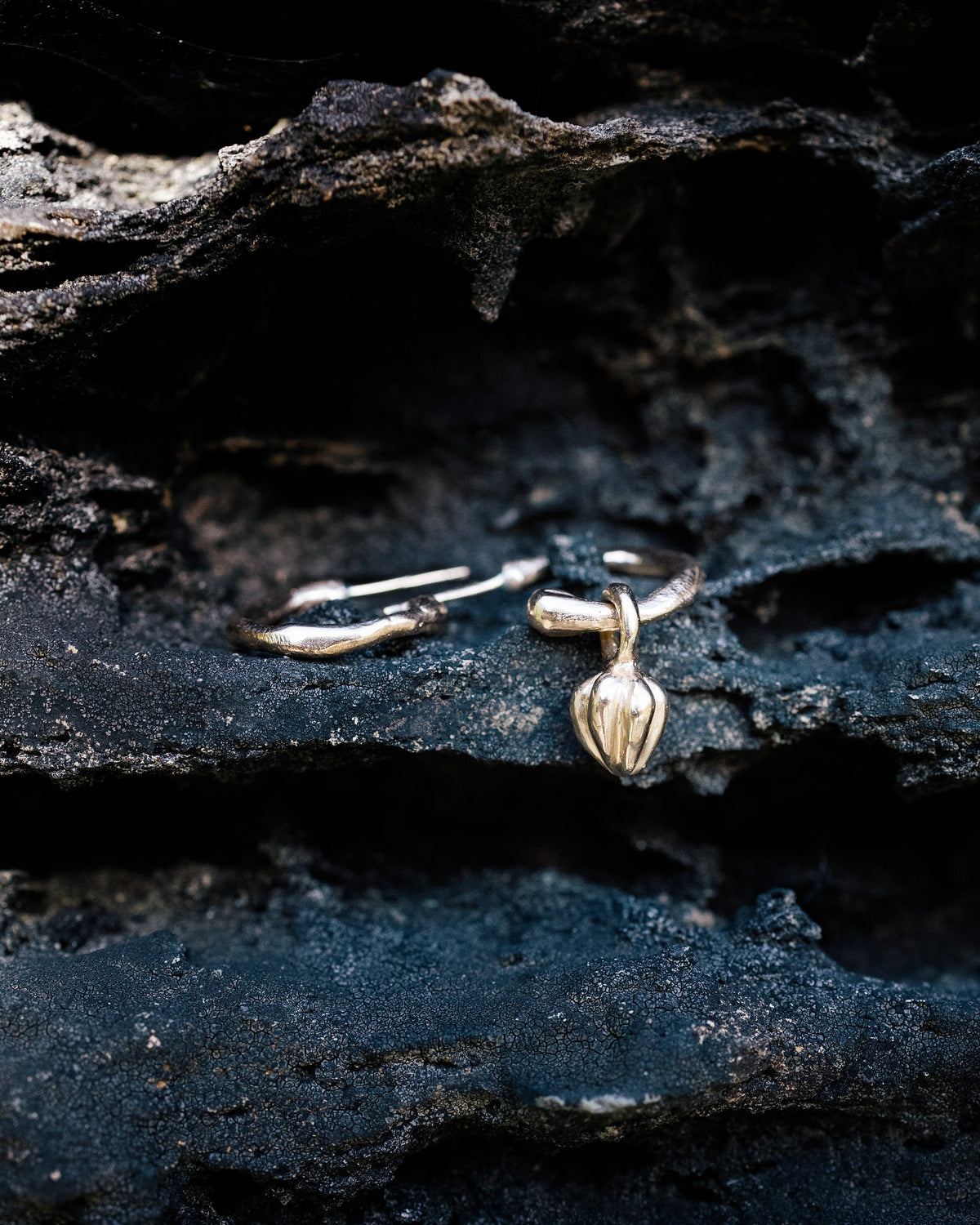 A pair of gold Migaru creole earrings on dark, layered rock, one with a seed-shaped charm and one plain.